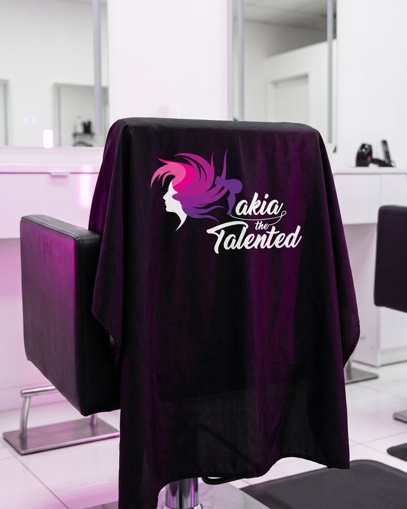 Mockup of the Takia the Talented logo printed in white on a black salon cape