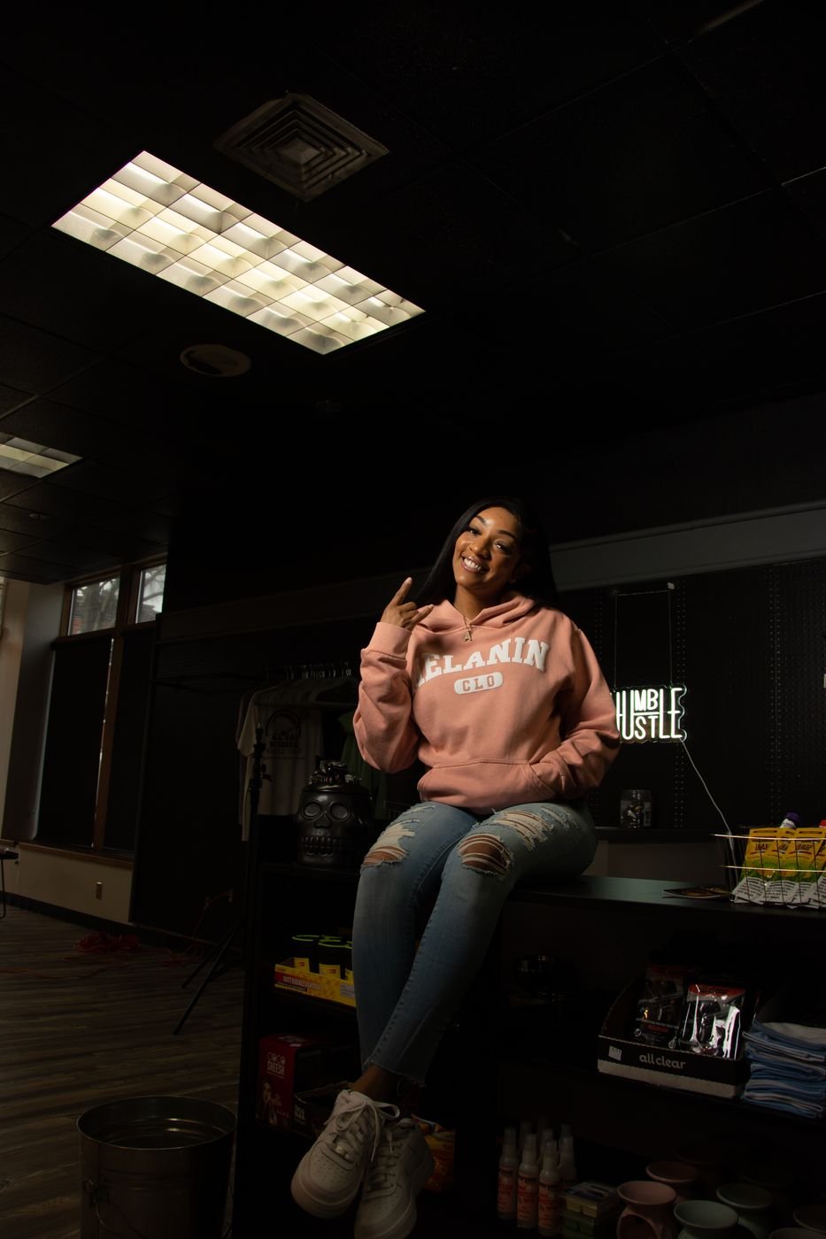Model in a blush Melanin CLO hoodie seated on a counter in a dark studio