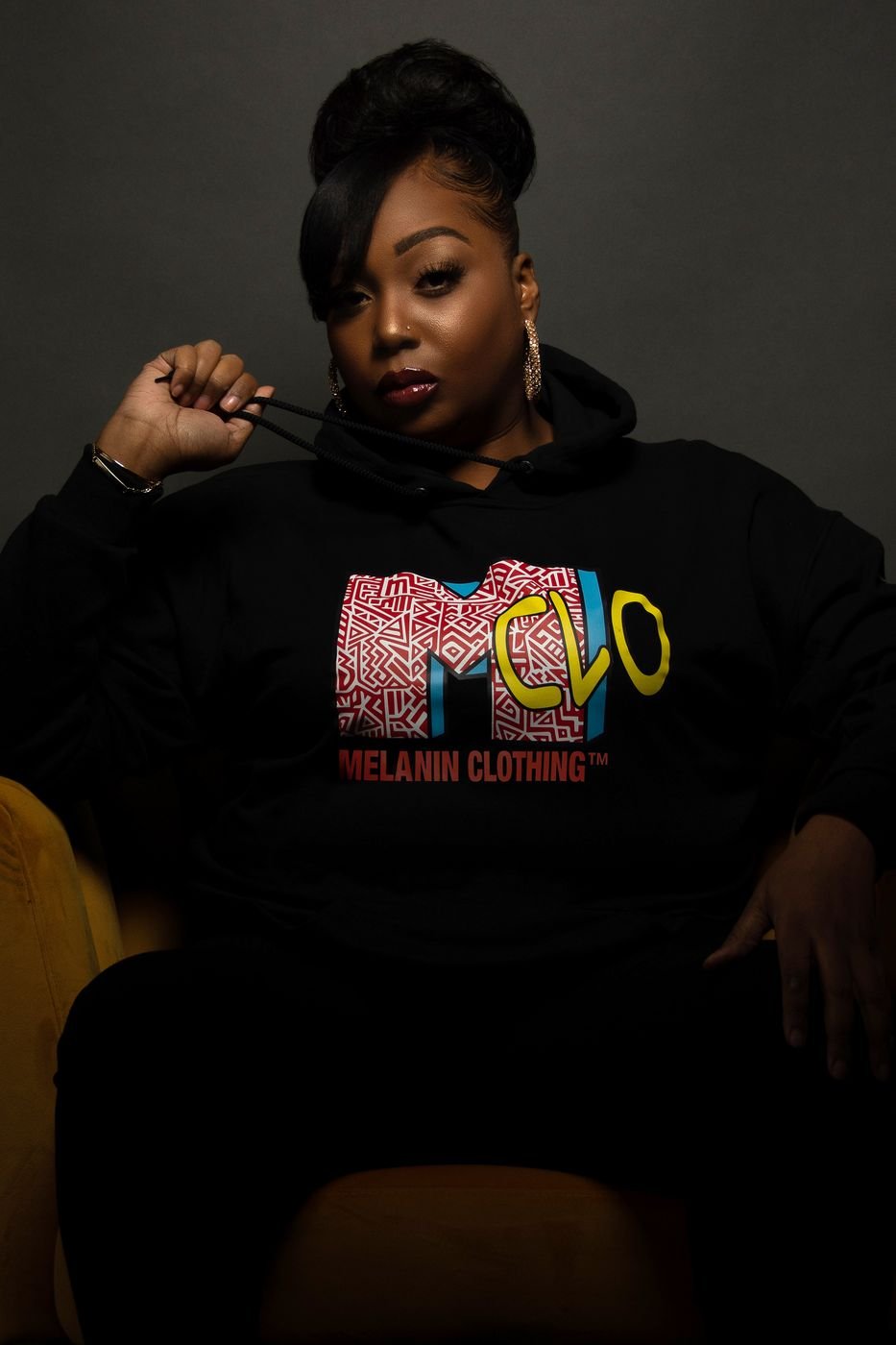 Model seated in a black Melanin Clothing graphic hoodie, studio portrait