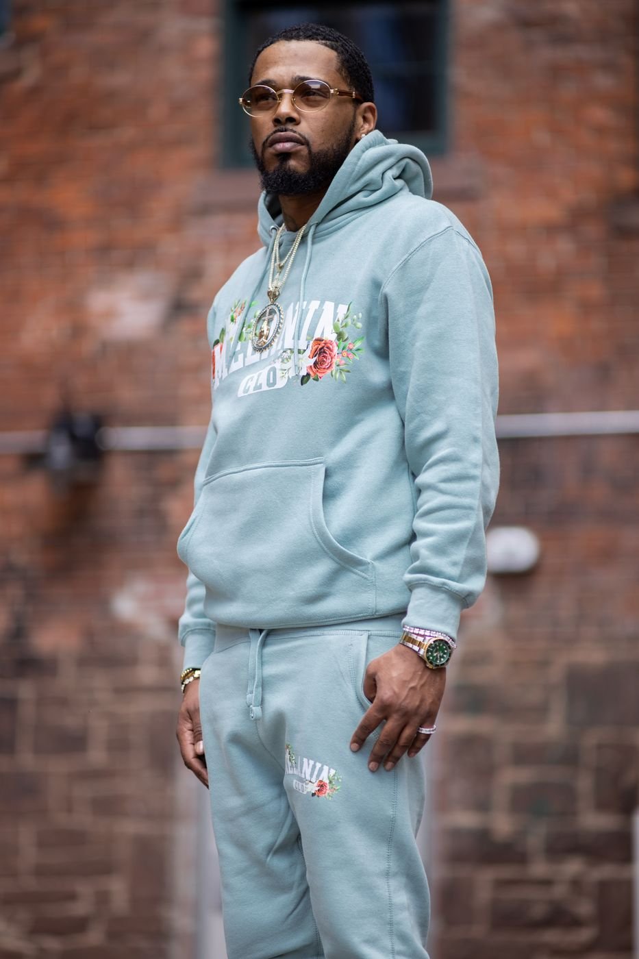 Full-length portrait of a model in a sage Melanin CLO tracksuit by a brick wall