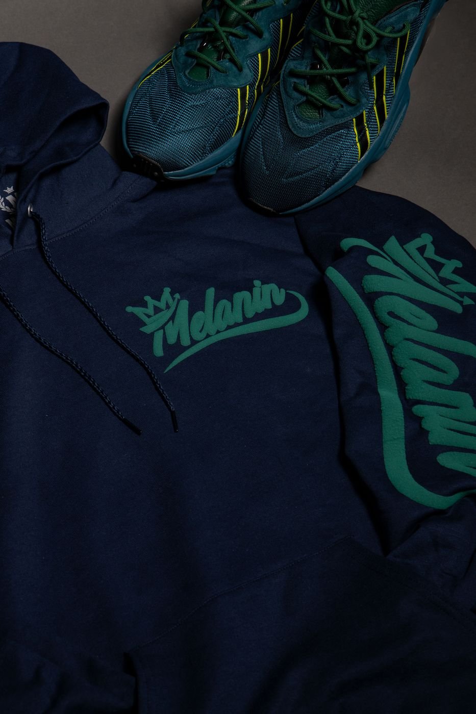 Flat lay of a navy Melanin hoodie with green script print and sneakers